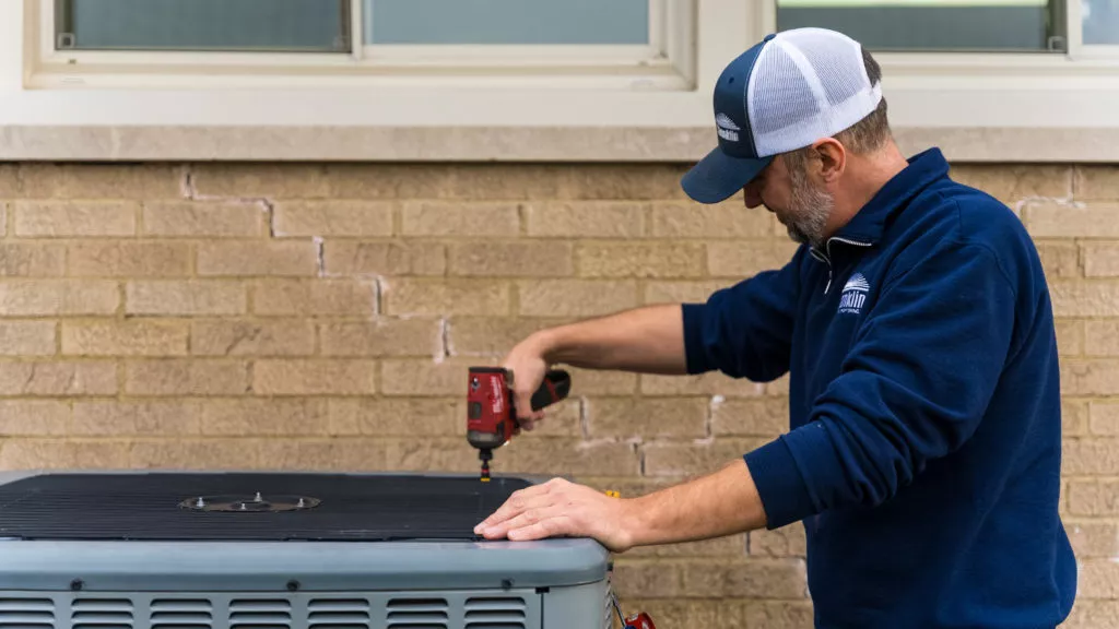 Technician performing AC maintenance on outdoor unit next to a tan brick home.